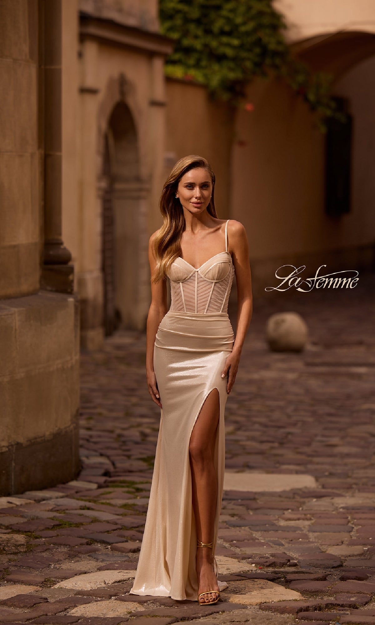 A woman wears the La Femme 32951 long prom dress, featuring a cream color, ruched bodice, and high leg slit, as she walks on a cobblestone street near stone buildings.
